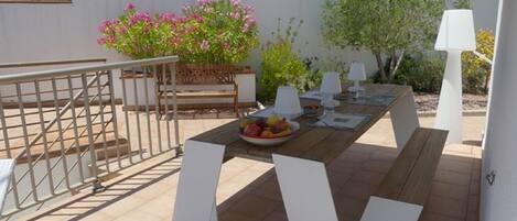 An outdoor patio with a wooden table set for dining, surrounded by white chairs.