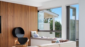A modern living room with a wooden panel wall, a round mirror, a sofa, and a window with a view of greenery. - Chic Beachside Abode, Sunrise Beach (Sunrise Beach)