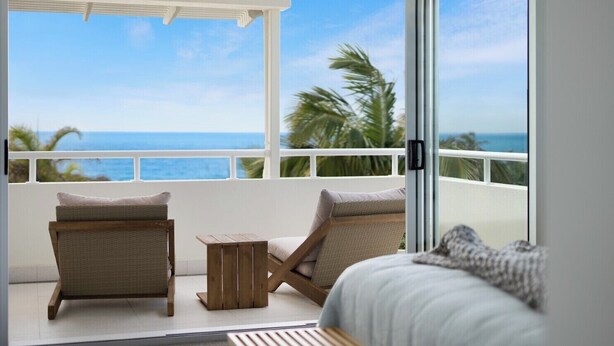 A balcony with a view of the ocean, two wicker chairs, a small wooden table, and a bed with a white headboard. - Chic Beachside Abode, Sunrise Beach (Sunrise Beach)