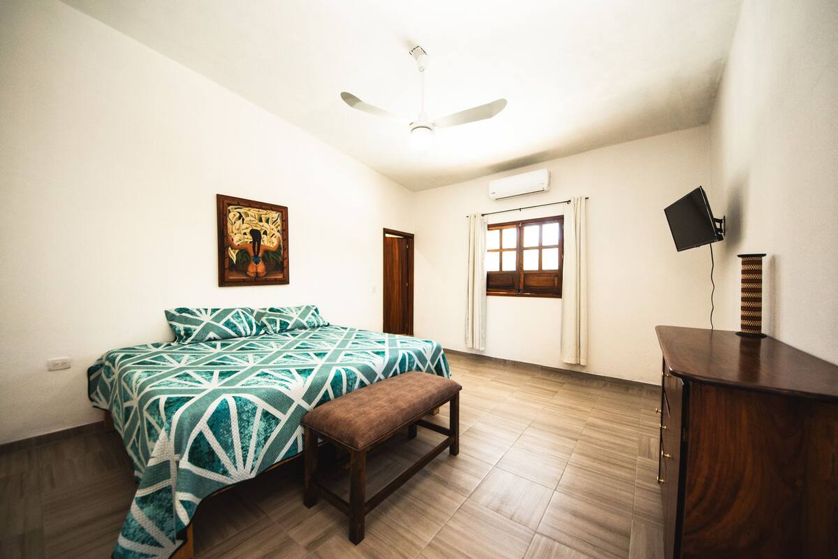 A bedroom with a bed, a bench, a ceiling fan, a window, and a wall-mounted TV.