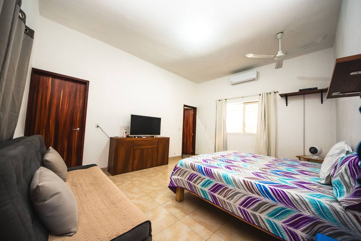 A bedroom with a bed, a TV on a wooden dresser, a ceiling fan, and a window with curtains.