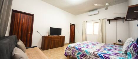 A bedroom with a bed, a TV on a wooden dresser, a ceiling fan, and a window with curtains.