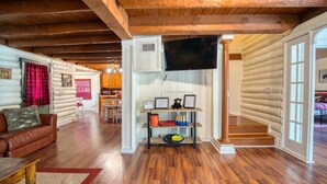 Interno di una baita in legno con soffitto in legno, una TV a schermo piatto montata a parete, una scala in legno e un’accogliente area salotto con un divano in pelle e un tavolino da caffè.