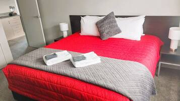 A hotel room with a red bedspread, a grey quilted bedspread, two towels, a grey pillow, and a white pillow.