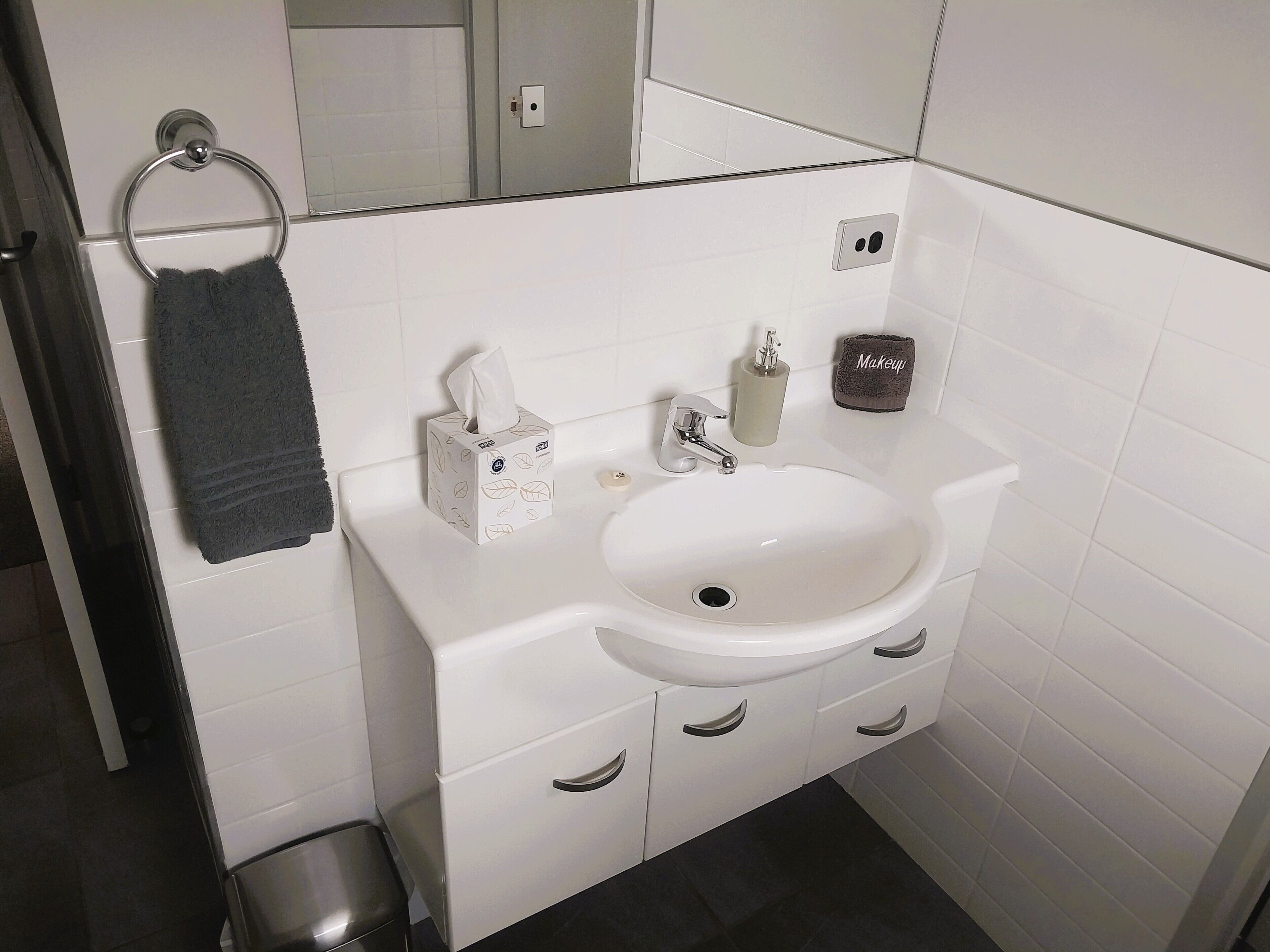 A bathroom with a white sink, a towel rack with a dark towel, a tissue box, a soap dispenser, and a makeup towel.