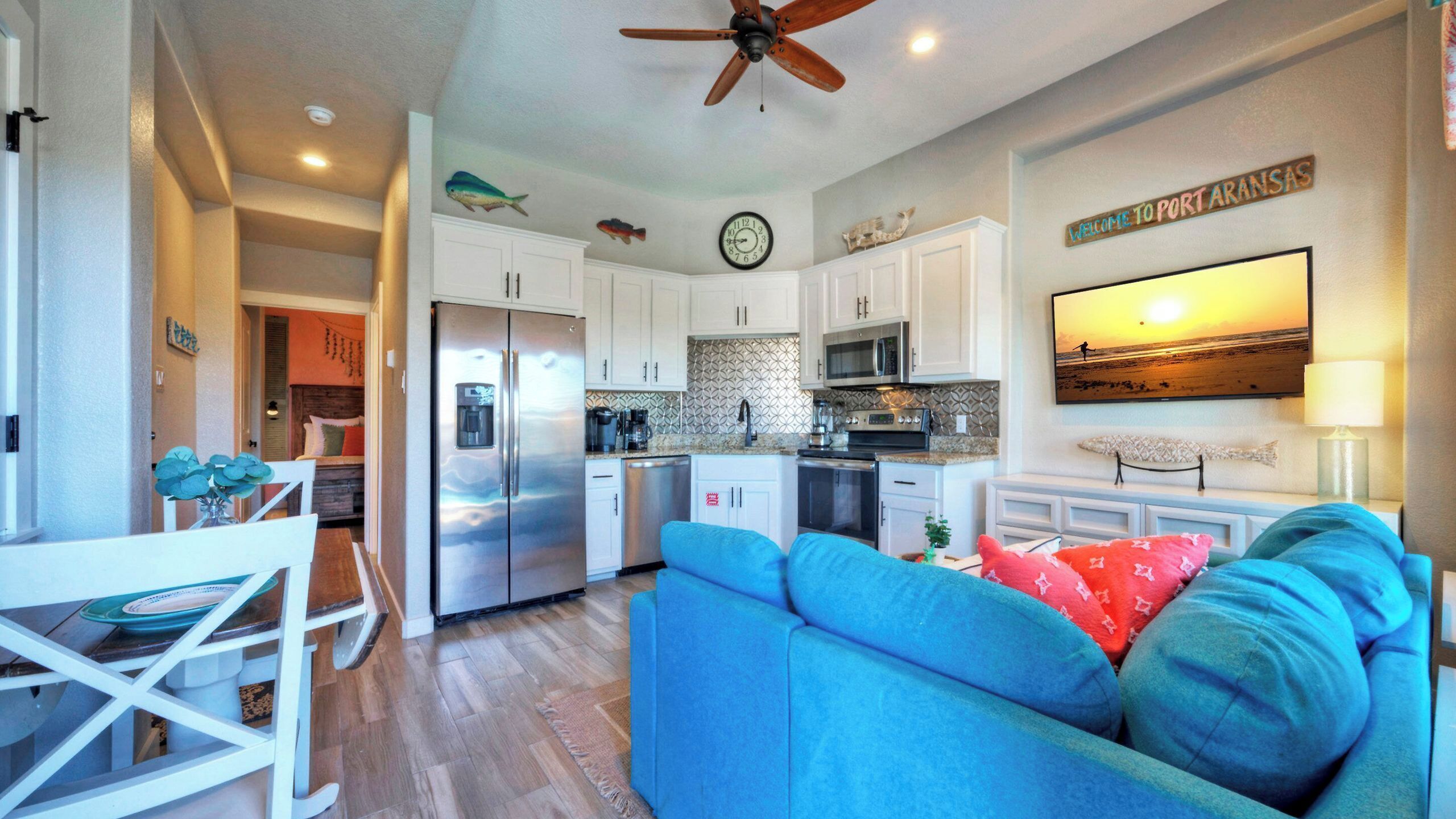 A modern kitchen with a refrigerator, microwave, and stove. There is a dining area with a table and chairs. The living room features a blue sofa and a television.