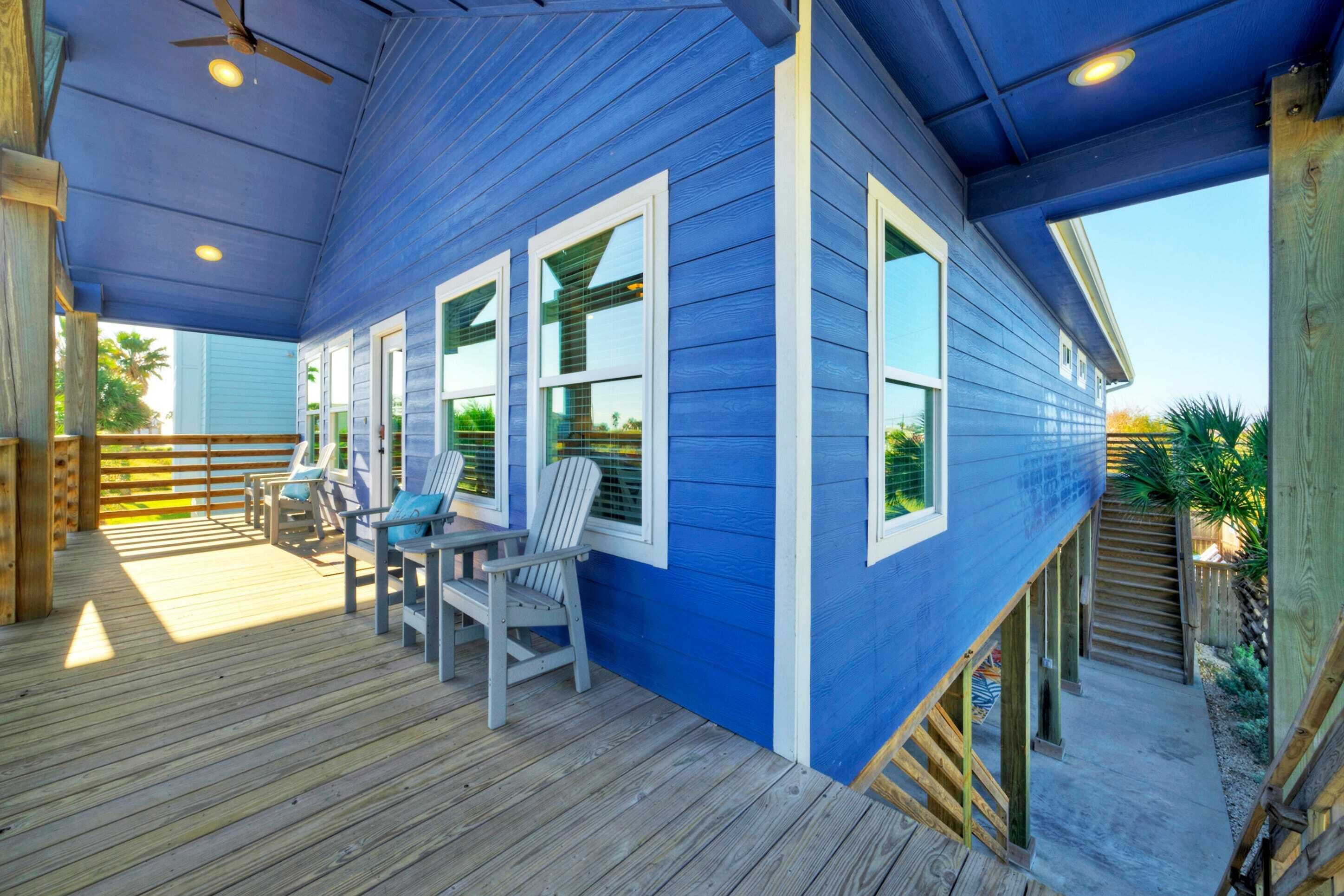 A covered deck with wooden flooring and Adirondack chairs, a blue house with white trim, and a wooden staircase leading to a lower level.