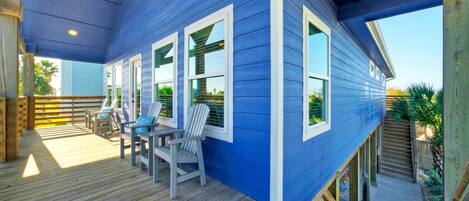 A covered deck with wooden flooring and Adirondack chairs, a blue house with white trim, and a wooden staircase leading to a lower level.