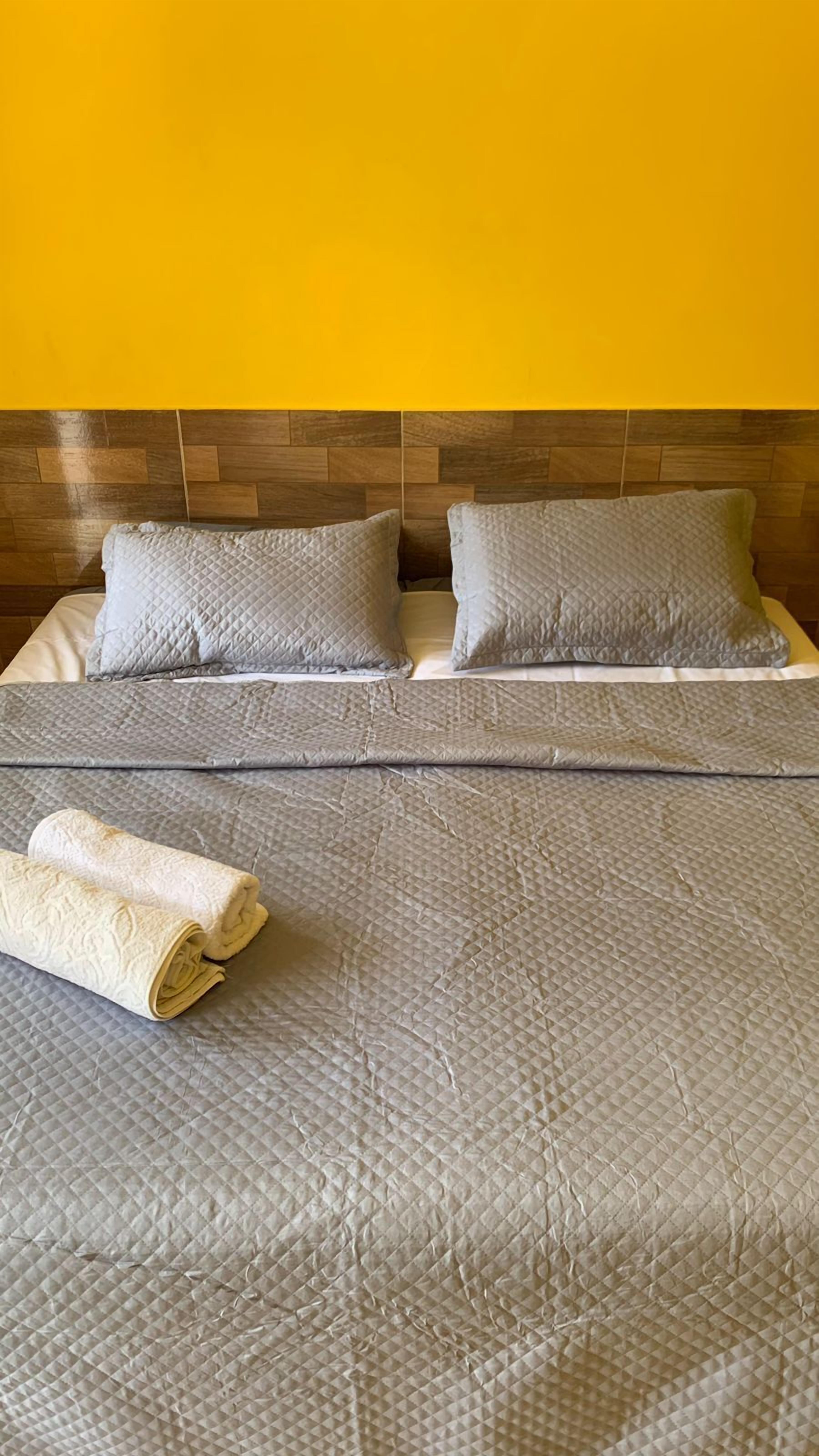 A double bed with a quilted grey bedspread and two pillows, a rolled white towel, and a wooden headboard against a yellow wall.