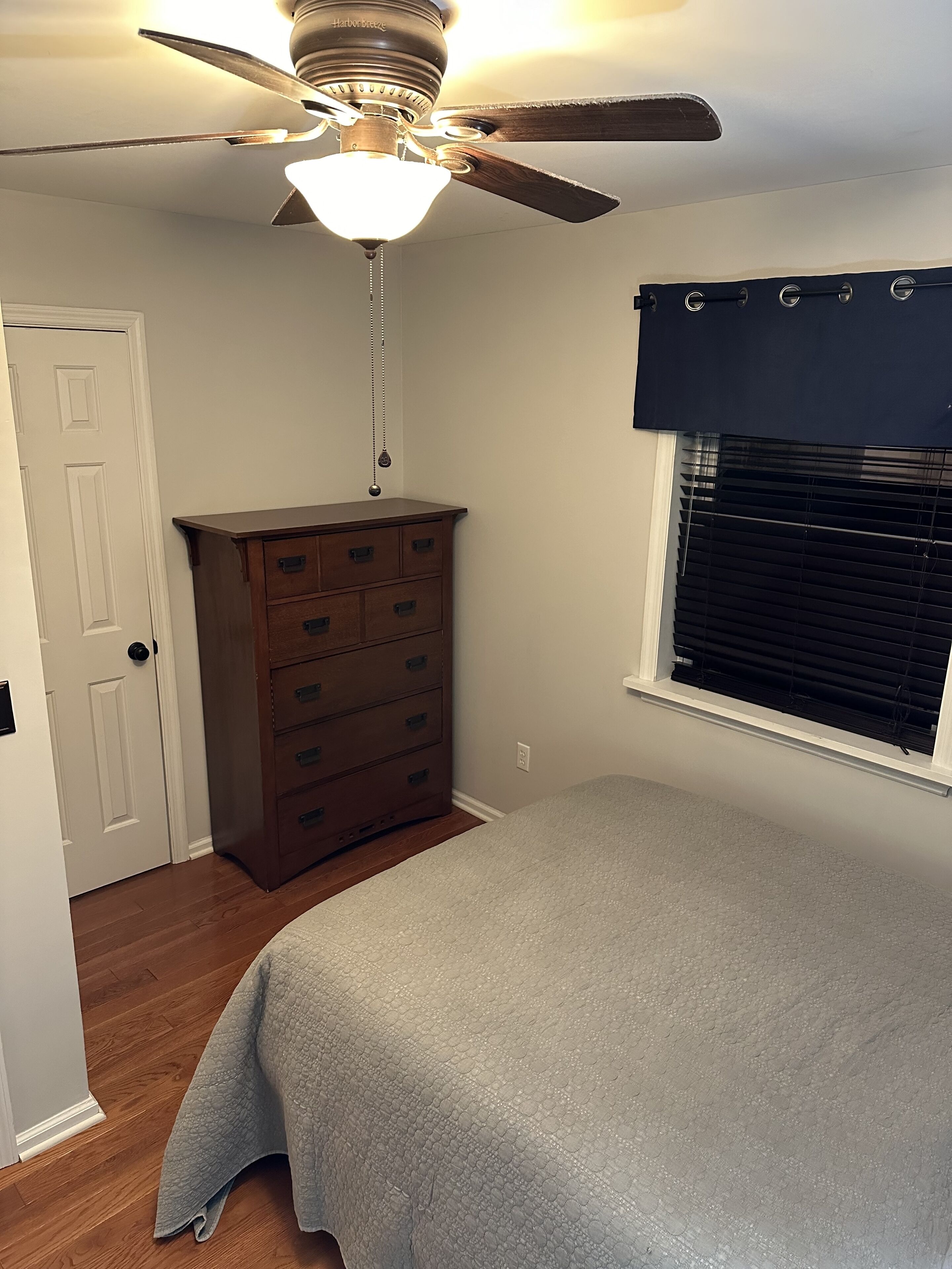 A bedroom with a ceiling fan, a dresser, a window with blinds, and a bed with a grey bedspread.