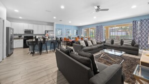 A modern living room with a sectional sofa, a dining area with a table and chairs, and a kitchen with white cabinets and appliances. - ⭐⭐⭐⭐⭐Spacious 10-BR Family Retreat Near Disney, Solterra Resort - Pool Spa, Game (Davenport)