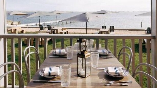 Une table à manger dressée avec des verres et des assiettes, donnant sur une plage parsemée de parasols.