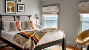 Un dormitorio con cama, una silla, un ventanal con vistas y cuadros en la pared.