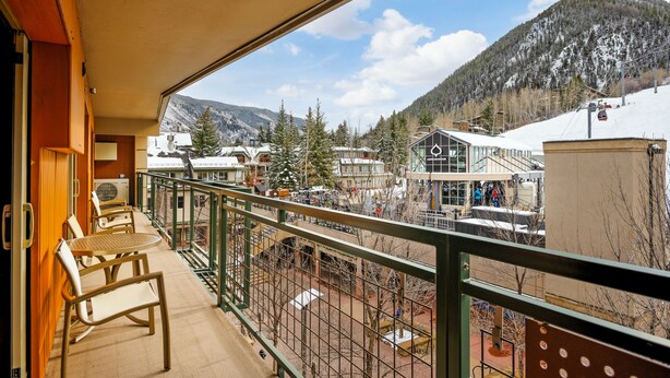 A balcony with a view of a snowy mountain, a building with a glass facade, and a chair by the railing. - North Of Nell, Unit 4b (Aspen)