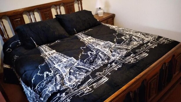 A bed with a black and white Eiffel Tower design bedspread, wooden headboard, and a bedside table with a lamp. - Hostal Gomero (Santa Cruz)