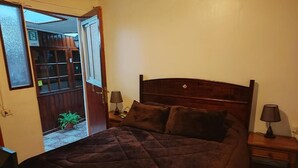 A bedroom with a large bed, two bedside tables with lamps, a wooden headboard, and a door leading to a balcony with a potted plant. - Hostal Gomero (Santa Cruz)