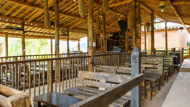 A rustic wooden structure with benches and tables, a brick wall, and a wooden ceiling with hanging lights. - Ciudadela el Artesano  (Carolina del Príncipe)