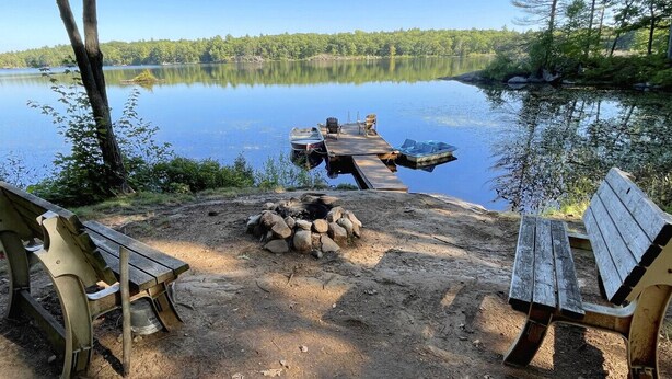 En campingplats vid sjön med en brygga, en eldstad och två bänkar.