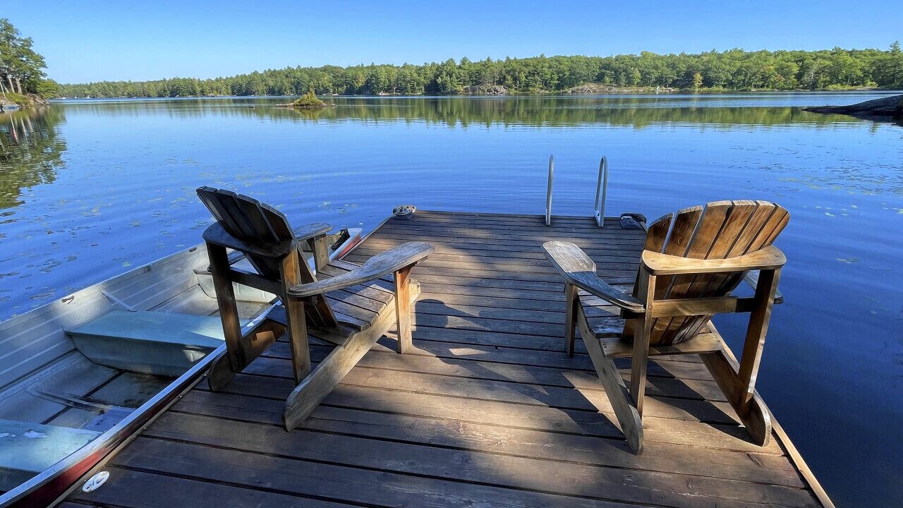 A wooden dock with two Adirondack chairs facing a calm lake, surrounded by trees.