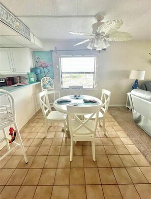 A compact kitchen with white cabinets, a dining table with four chairs, a ceiling fan, and a window with blinds. - Ponce Inlet 6th Floor Ocean View Corner Unit- 2bdrm 2bath- Non Drive Beach  (Ponce Inlet)