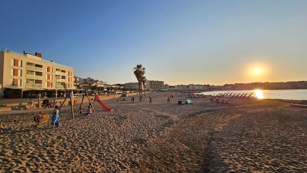 A beach with a playground, palm trees, and a building with balconies. - Casa Sebastian (L'Escala)