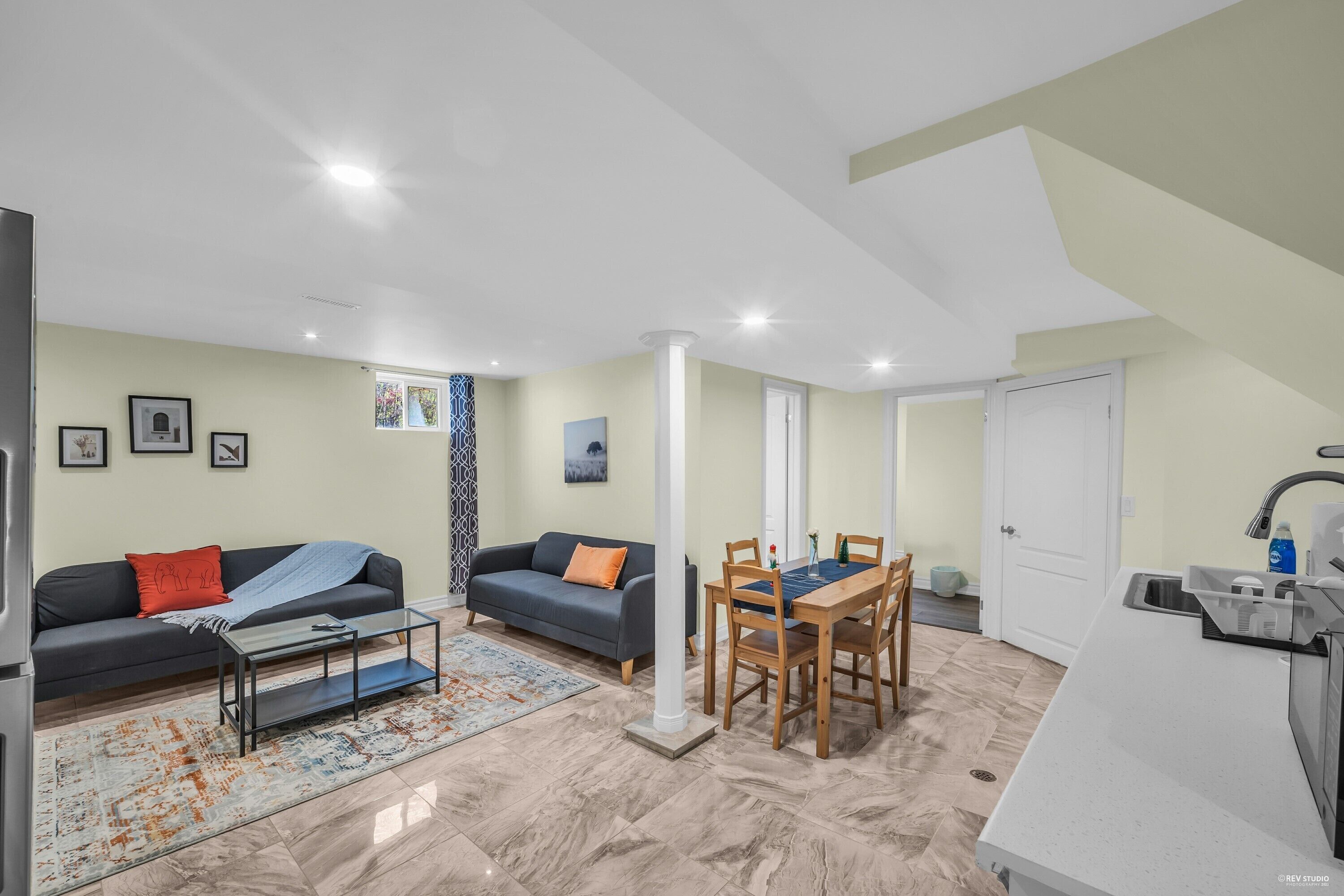 A modern living room with a sofa, coffee table, and dining table. There is a kitchen area with a sink and cabinets.