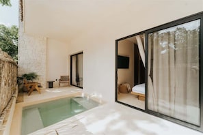 A modern outdoor pool area with a stone wall, wooden furniture, and a glass door leading to a bedroom. - Casa Coba Plunge Pool Beach Club Aldea Zama (Tulum)