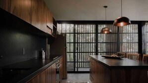 A modern kitchen with dark wood cabinets, a central island, pendant lighting, and a view through a large window with vertical blinds. - Jungle Modern Loft 101 Aldea Zama Walk Everywhere (Tulum)