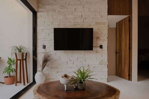 Une pièce avec un mur en briques, un téléviseur fixé au mur, une table ronde en bois ornée de plantes et une porte donnant sur une autre pièce.