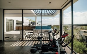 A modern room with a large window, exercise equipment, a pool, and outdoor seating.