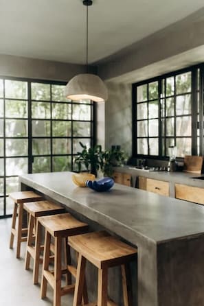 A modern kitchen with a long island, wooden stools, and a hanging light fixture.