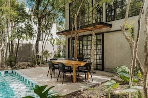 A modern outdoor dining area with a pool, surrounded by trees and plants.