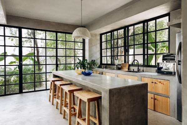 A modern kitchen with large windows, a central island with stools, and a hanging light fixture.