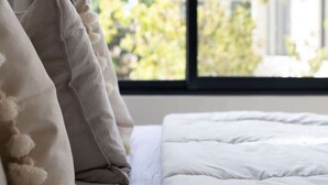 A bed with white bedding and fluffy pillows, a window with a view of buildings and greenery, and a curtain partially drawn. - Casa Kaatal Sunny Penthouse Pool Beach Club Access (Tulum)