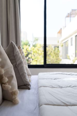 A bed with white bedding and fluffy pillows, a window with a view of buildings and greenery, and a curtain partially drawn. - Casa Kaatal Sunny Penthouse Pool Beach Club Access (Tulum)