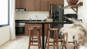 A modern kitchen with wooden cabinets, a black countertop, a stainless steel refrigerator, and two wooden stools. - Casa Kaatal Sunny Penthouse Pool Beach Club Access (Tulum)