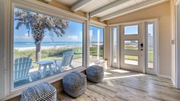 A spacious room with large windows overlooking a beach, wooden flooring, and a door leading outside.