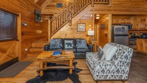 Interior de una cabaña de troncos con una escalera de madera, un sofá de cuero negro con un cojín de oso, un sofá estampado, una mesa de centro de madera y una zona de cocina con refrigerador y estufa.
