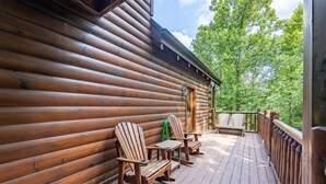 Una terraza de madera con sillas Adirondack y una mesa, rodeada por una cabaña de madera con un porche cubierto.