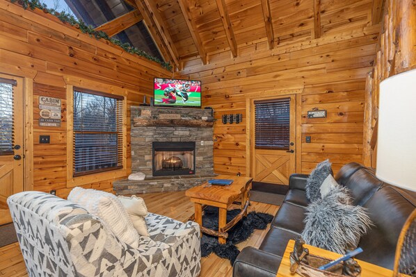 A cozy cabin interior with a stone fireplace, wooden walls, a television mounted above it, and a comfortable seating area with a couch and chairs. - A Bear On The Ridge-Gatlinburg, Pet Friendly (GATLINBURG)