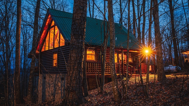 A cabin with a green roof, surrounded by trees, and lit by warm interior lights. - A Bear's View-Gatlinburg, Resort Setting, Sleeps 6 (GATLINBURG)