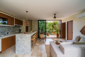 A modern kitchen with a marble island, wooden cabinets, and a dining area with chairs. There is a sliding glass door leading to a balcony with potted plants.