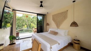 A bedroom with a large bed, a desk, a chair, a hanging light fixture, and a view of greenery outside.