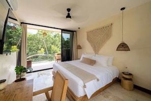 A bedroom with a large bed, a desk, a chair, a hanging light fixture, and a view of greenery outside.