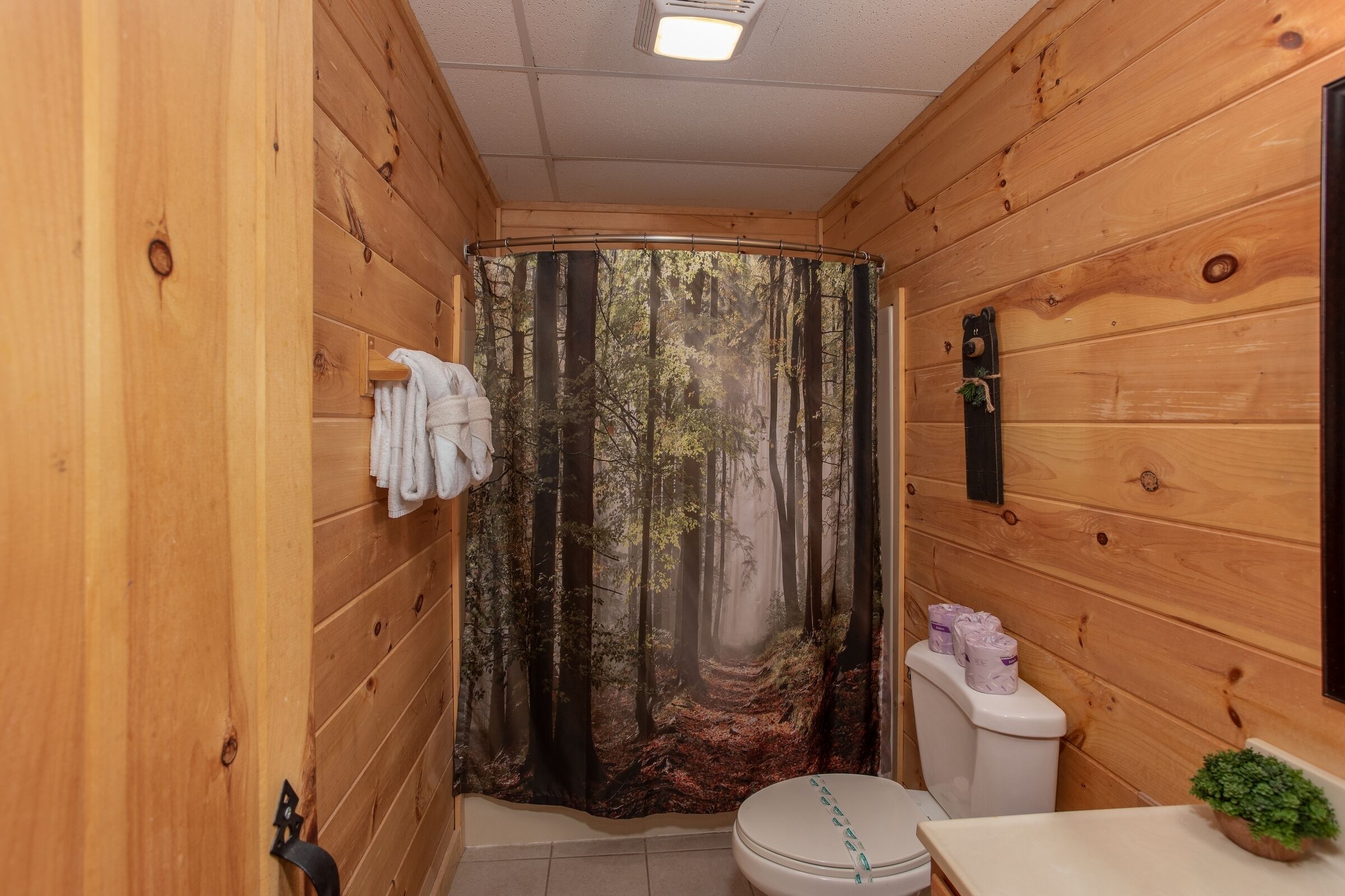 A wooden cabin bathroom with a forest-themed shower curtain, a toilet, and a sink with a plant.