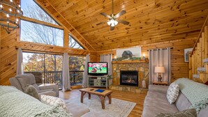 A cozy cabin interior with a wooden ceiling, a television, a fireplace, and a large window overlooking a forest.