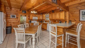 A wooden cabin interior with a large dining table, white chairs, wooden cabinets, a kitchen area, and a fireplace. - Game Room*Families Welcome*Large Hot Tub*Easy Access Roads (PIGEON FORGE)