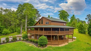 A two-story log cabin with a balcony, surrounded by a well-maintained lawn and trees. - Game Room*Families Welcome*Large Hot Tub*Easy Access Roads (PIGEON FORGE)