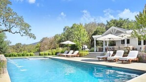 Una piscina con sillas de descanso y sombrillas, rodeada de vegetación y un edificio con un porche cubierto.