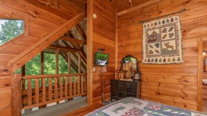 A cozy cabin with a wooden interior, a loft bedroom with a view, a quilted wall hanging, and a small wooden cabinet.